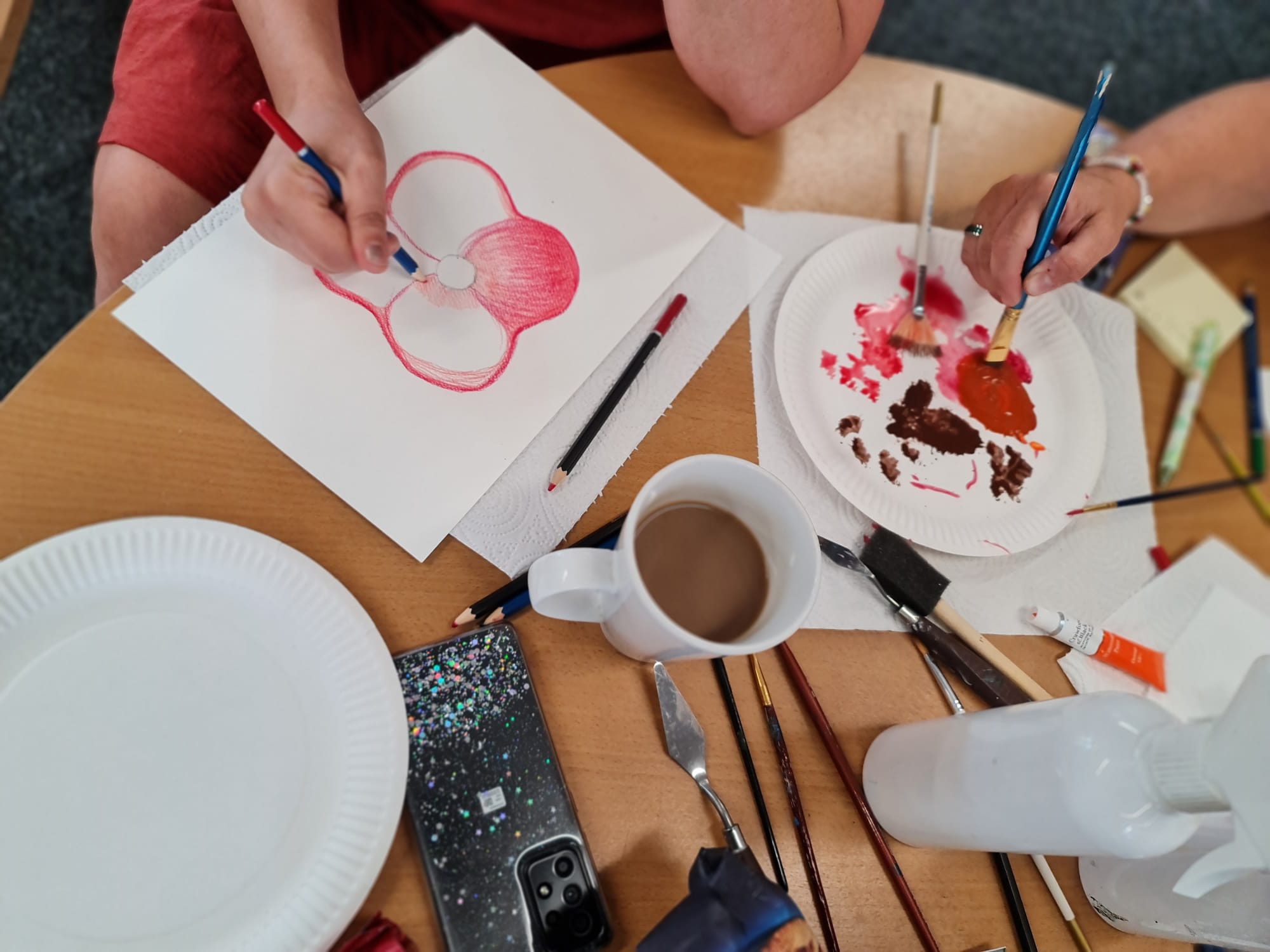 Table with art supplies and hands of two people, one painting a red poppy